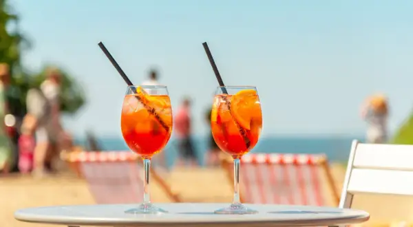 Dwa kieliszki Aperol Spritz z pomarańczą i słomką na stole, w tle plaża i morze.