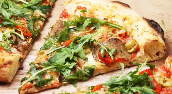 Kawałki pizzy z rukolą, oliwkami i warzywami na drewnianym tle.